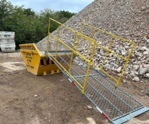 Caçamba Multilix amarela com rampa de acesso em tela metálica e corrimão amarelo, posicionada em canteiro de obras ao lado de grande pilha de entulho.
