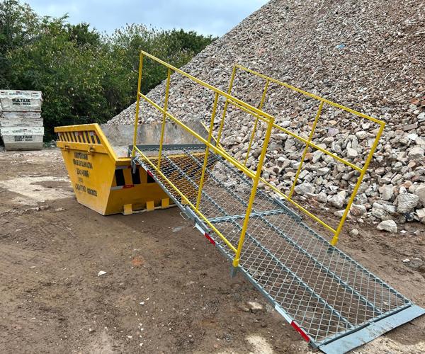 Home 84 Caçamba Multilix amarela com rampa de acesso em tela metálica e corrimão amarelo, posicionada em canteiro de obras ao lado de grande pilha de entulho.